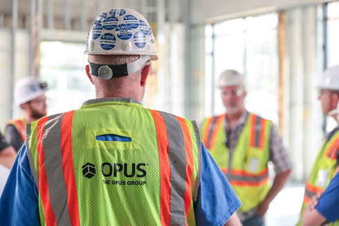 back of man wearing hardhat and safety vest with the words "The Opus Group" in the foreground and three men wearing hardhats and safety vests in background