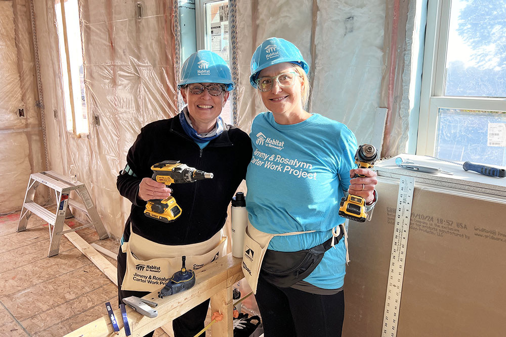 two women hold power drills and smile for a photo inside a home under construction