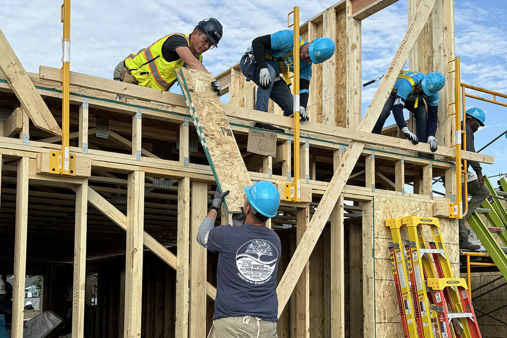 a man with his back to the viewer hands a piece of particle board up to another man on the second floor of a home under construction