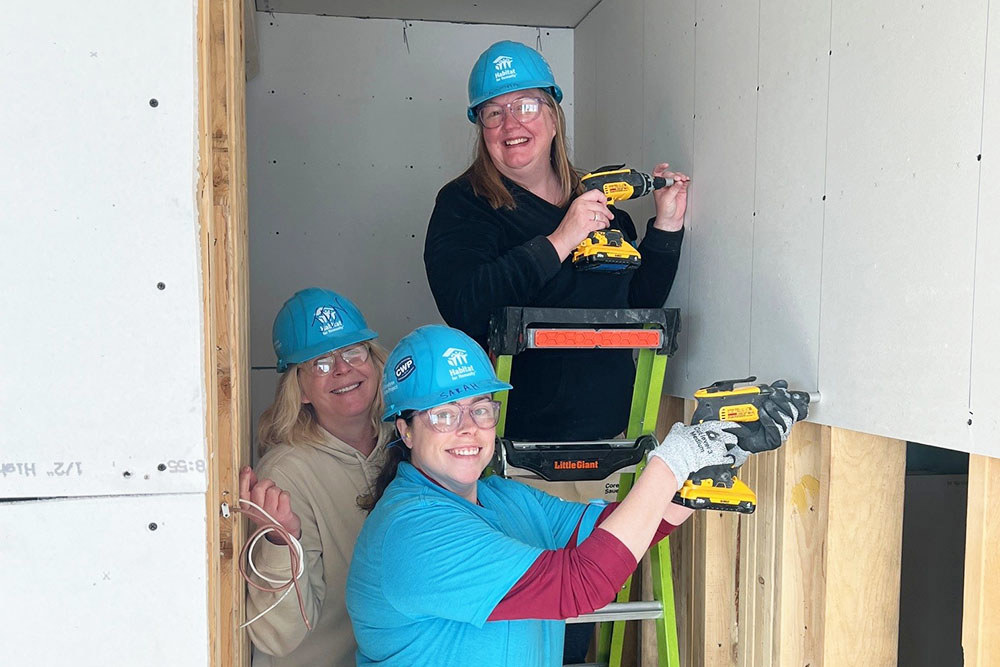 3 smiling women in hard hats stand inside a niche in a home under construction