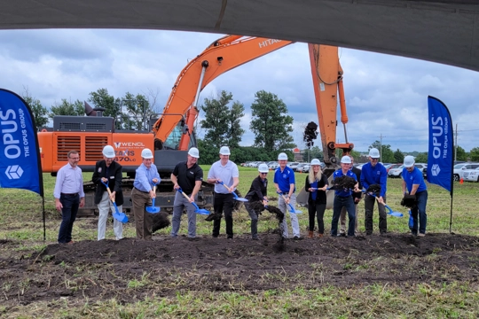 a group of people stand in a line holding shovels above a pile of dirt on the ground with construction equipment in the background