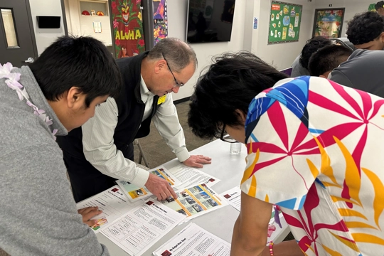 man and two teenagers looking at papers on table in classroom