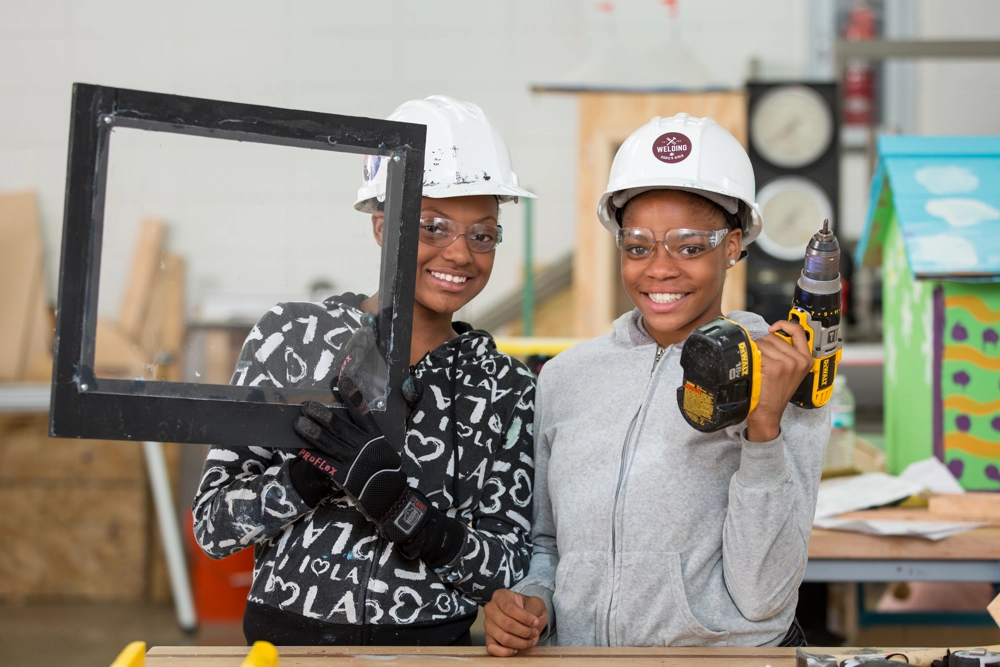 two girls in hardhats with one holding a drill and the other holding a black picture frame