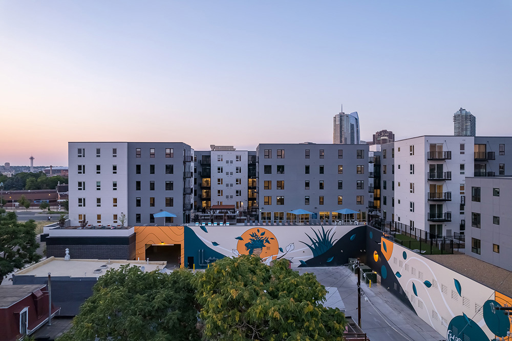 aerial view of multifamily development with colorful murals on ground level and rooftop amenity space in front of apartment towers