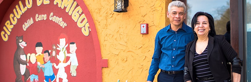 A Hispanic couple stands in front of the entrance to their child care center business.