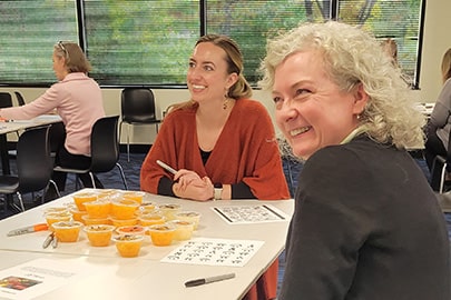 Two smiling women looking at something off camera sit at a table with snacks and craft supplies.