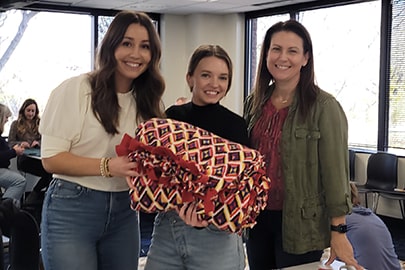 Three smiling women hold a folded fleece tied blanket.