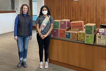 A woman and a girl stand in an office lobby with boxes of Girl Scout cookies.