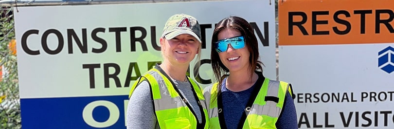 Two young women with high visibility yellow construction vests stand in front of signage at a construction sit.