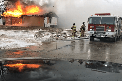 A fire truck and two firefighters observe a building on fire during a training session.