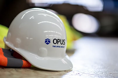 A hard hat with The Opus Group on the front sits on top of a high visibility construction vest.