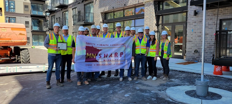 A group of people in construction personal protective equipment PPE including bright vests and hard hats stand in front of a building holding a banner.