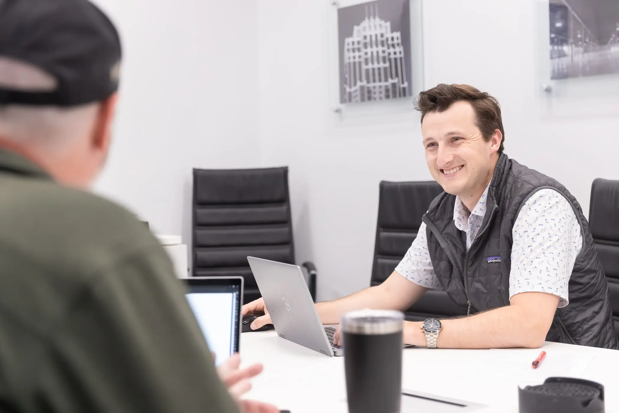 a smiling man sits at a conference room table with a laptop in front of him across from another man.